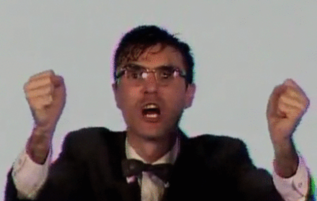From the Talking Heads 'Once in a lifetime' music video: A man in a jacket, shirt and bowtie, with fists raised as if in frustration