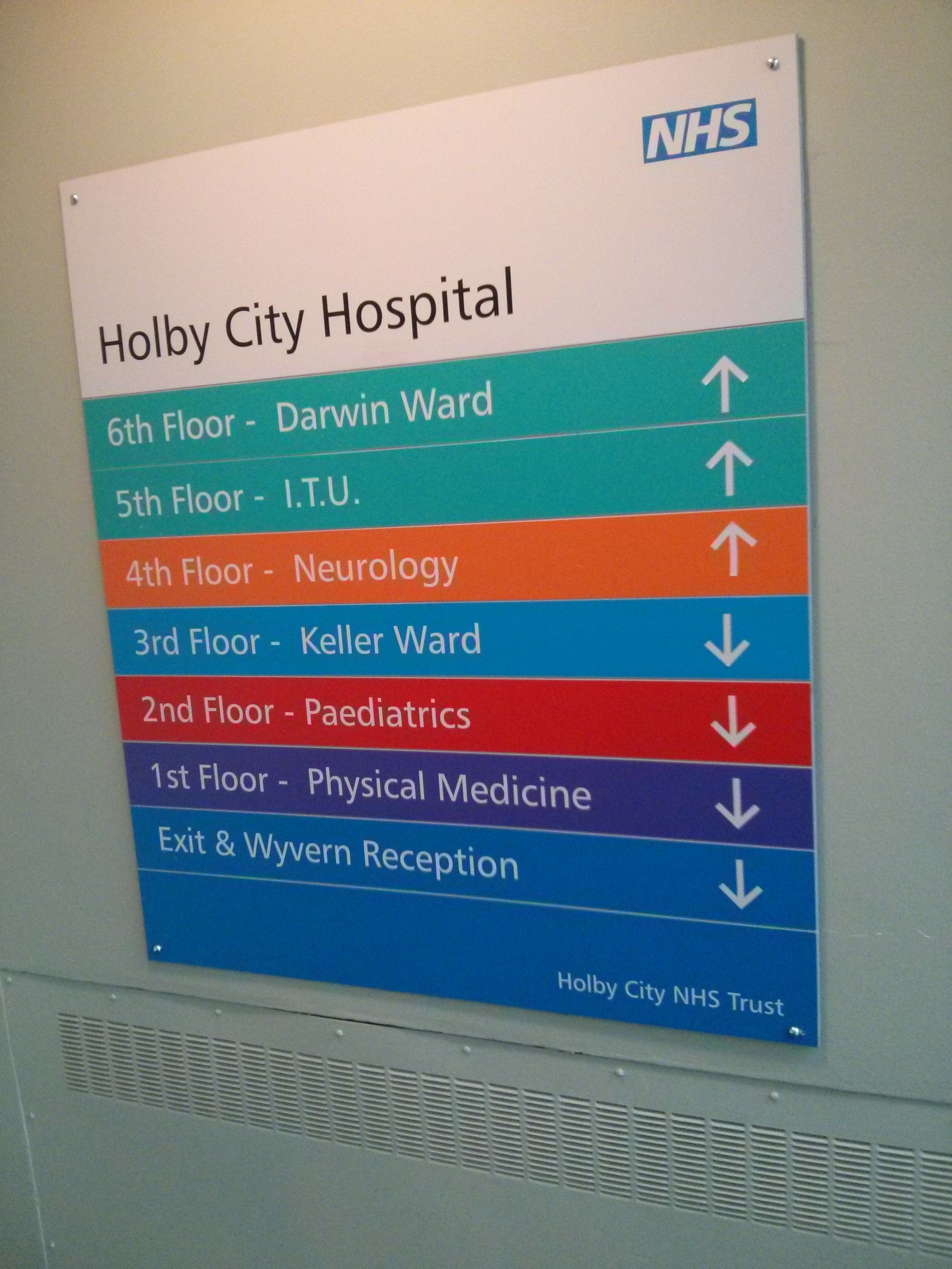 A sign on the wall in a stairwall of a normal office building, somewhere on the BBC Elstree site. The sign from 'Holby City NHS Trust', and lists what departments are on each floor: Neurology, Keller Ward, Paediatrics, etc. To explain: Holby is a fictitious hospital drama series; Holby City Hospital does not exist.