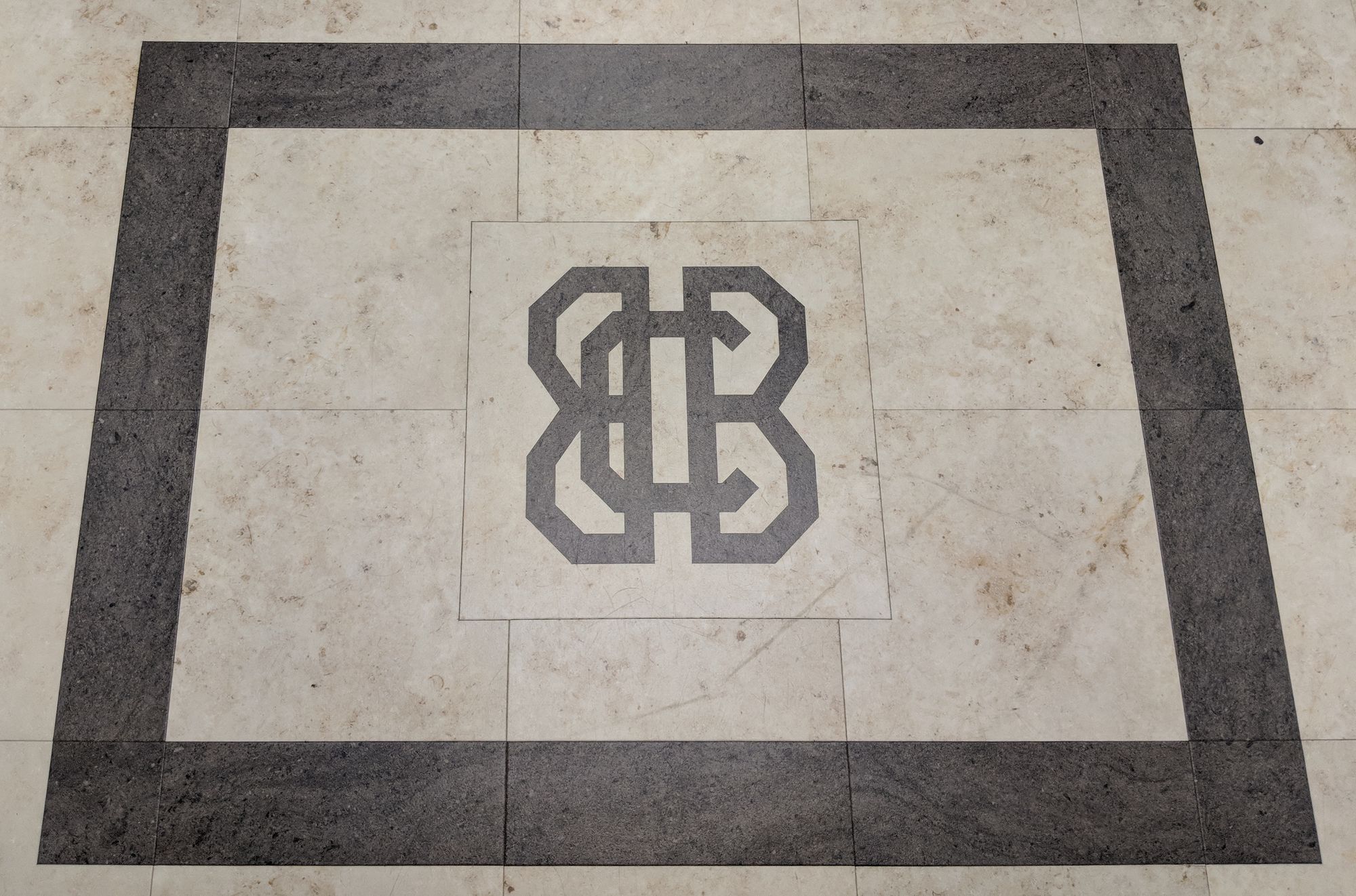 A marble-pattern tiled floor with light beige square tiles. Central, and on its own special tile, is a monogram of the letters B,B,C in an art deco style. Surrounding the whole thing is a thick dark border.