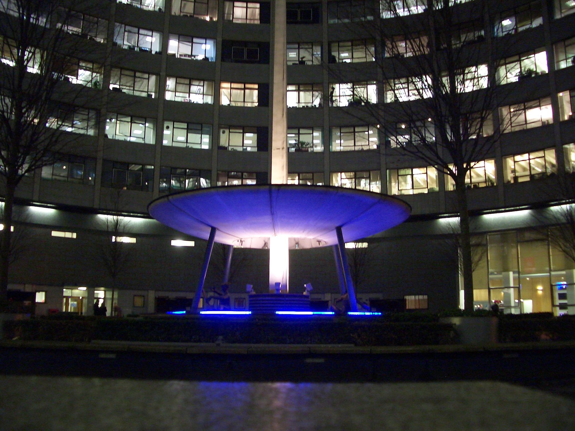 The iconic fountain at night, uplit in blue. In the background, the tall, curving wall of the surrounding building.