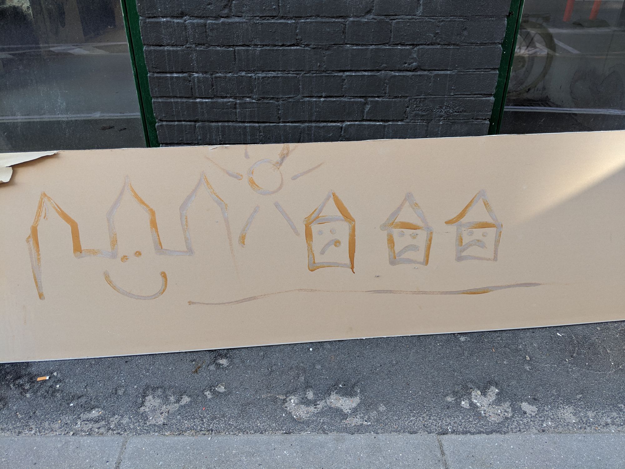 Lying on a ground, a piece of cardboard? wood? with a simple picture crudely painted on it: the sun, and on the left, what looks like a castle, smiling; and on the right, three small houses, who are sad.