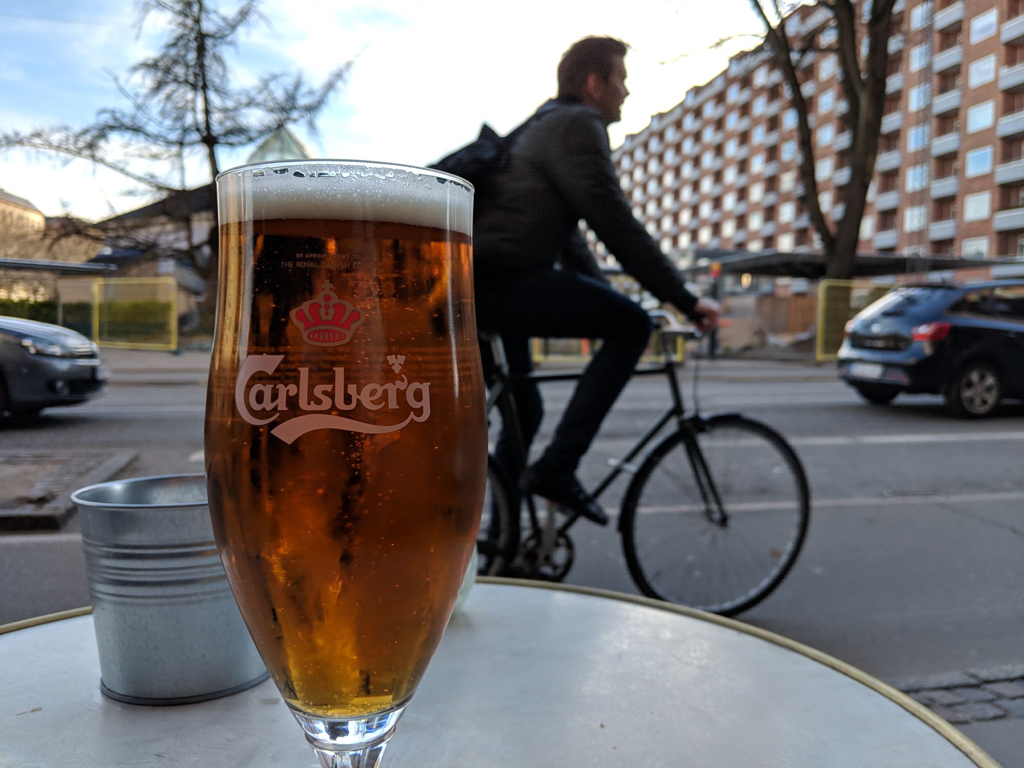 Close-up of a glass of Carlsberg beer on a table, outside a cafe. In the background, blurred (but only a couple of metres away), a man cycles past in the bike lane. Beyond that is a road, a couple of cars, and an apartment building on the right.