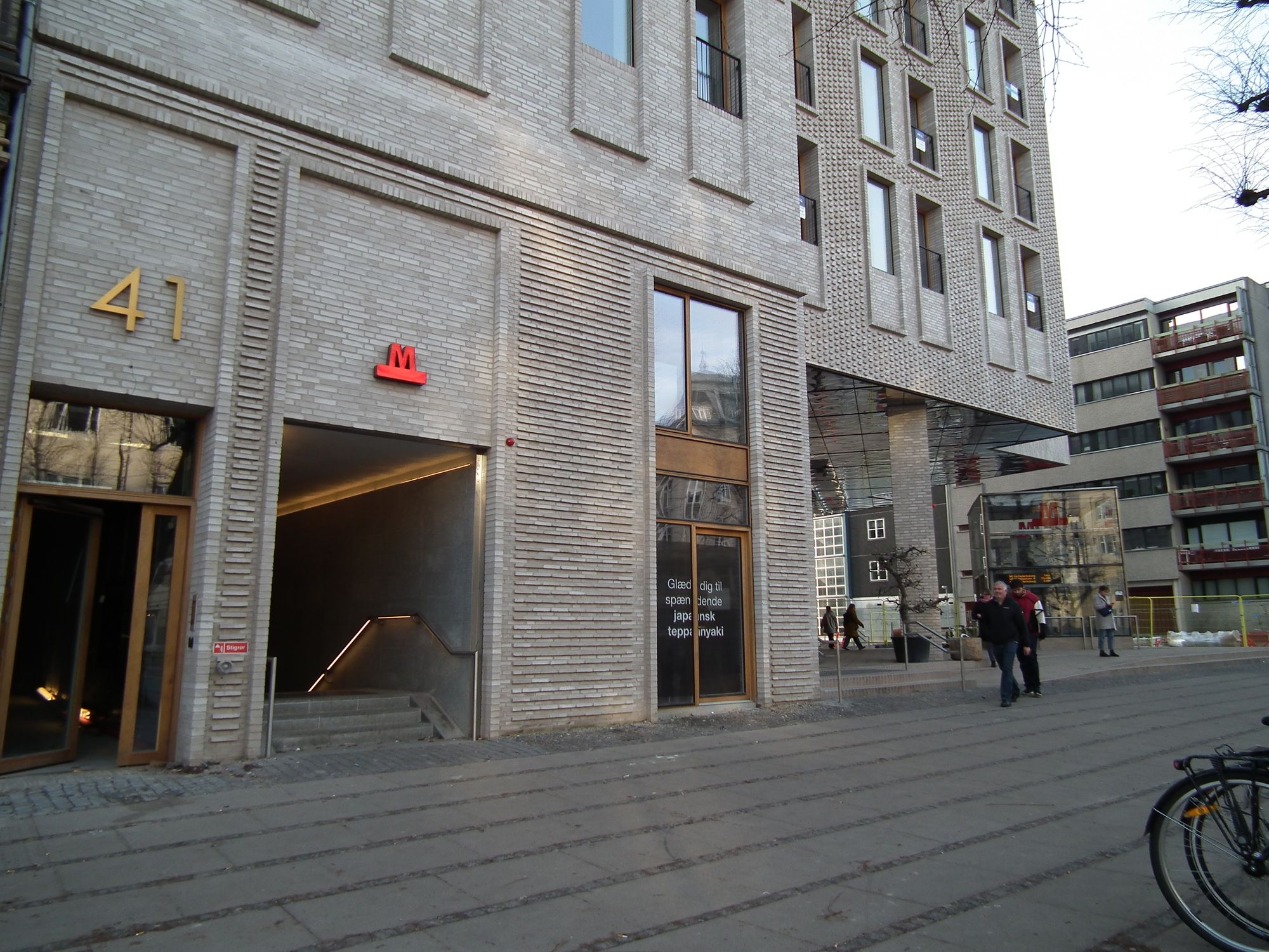 a modern building, at least 4 storeys tall (we can't see the top), next to a pavement. The corner part of the ground floor is 'cut out', the building above supported by columns. There is a glass elevator head here.
