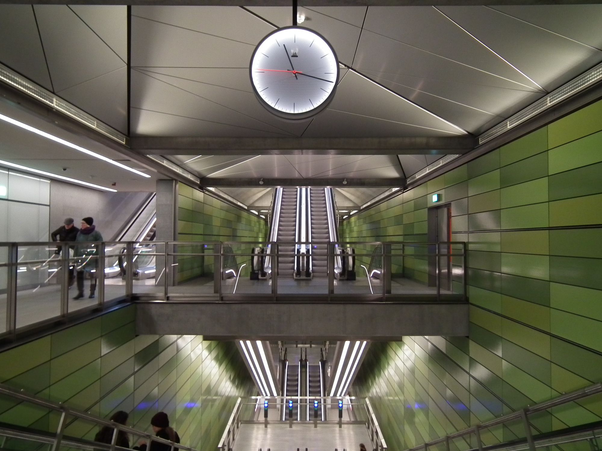 The inside of the station. The two large side walls are covered in rectangular tiles of various shades of green. There is white lighting, sometimes in plain sight, sometimes concealed.