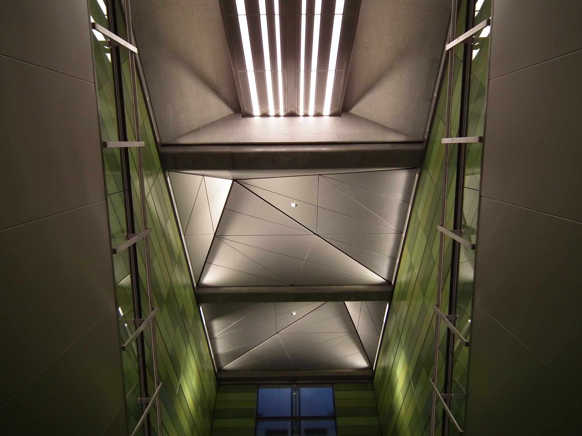 Looking up to the roof. Three square sections, one of which has simple white strip lights, where the other two rise into skew pyramids, composed of tesellating triangular tiles, and concealed uplighting.