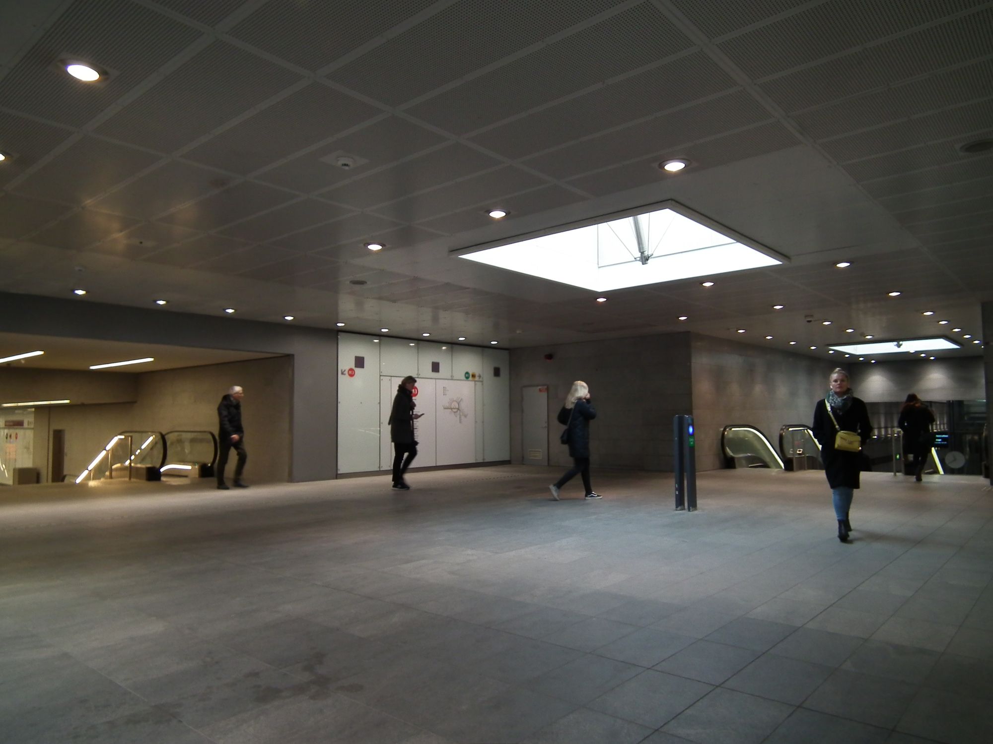 A modern and clean station interchange area, maybe 10 metres square. A (genuine) skylight provides natural illumination. A few people are here, going on their way.
