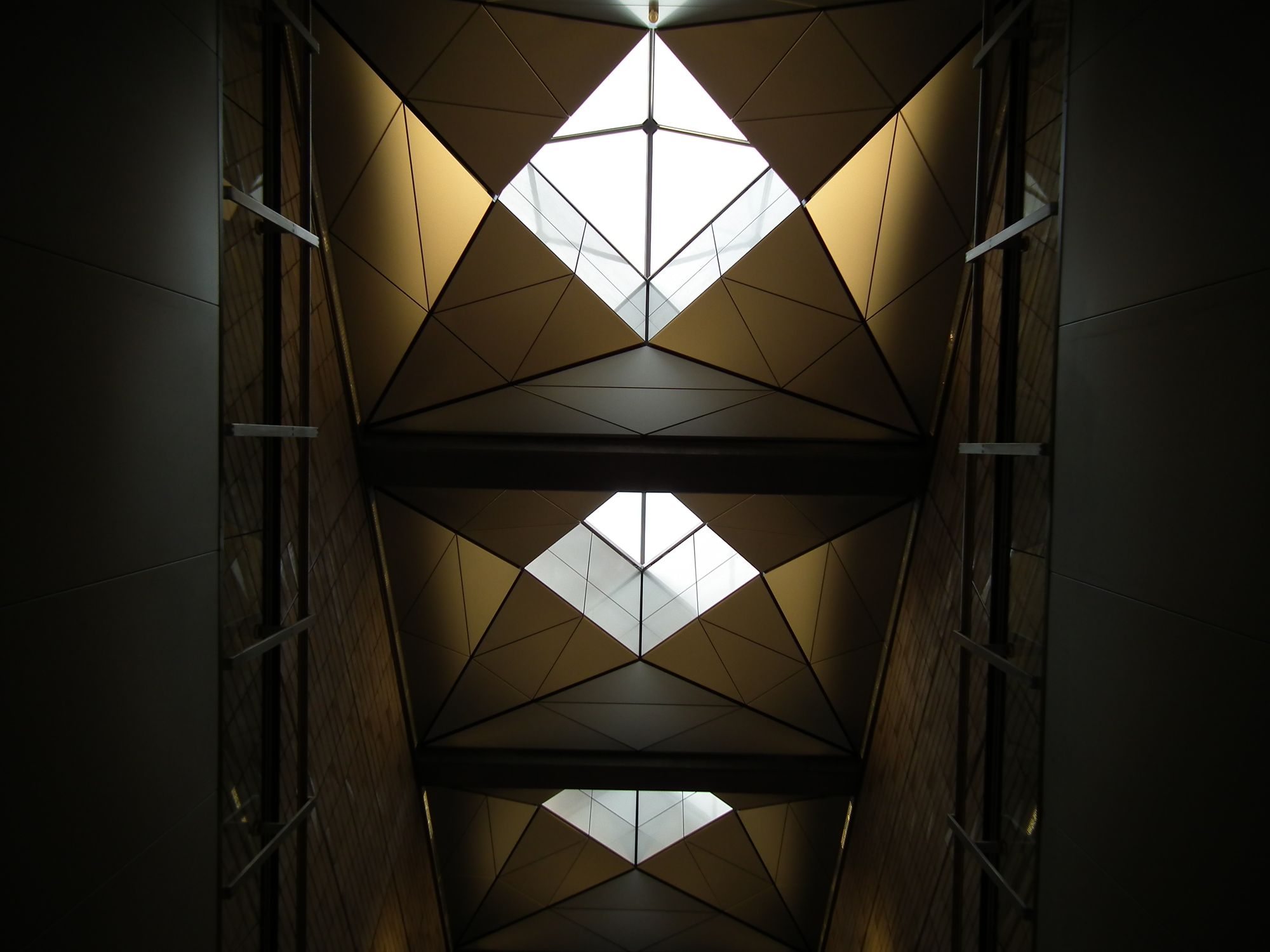 Looking up to the three skylights. Each is square, with a centre which is also square (but offset 45 degrees) and rises to allow sunlight in. Around the sunlight centres, there are 8 gold-brown triangles, like petals.