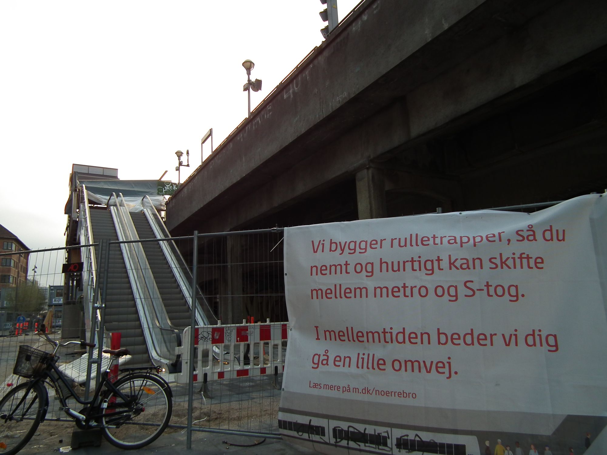 Outside, street level, showing the S-train line interchange, with escalators. However, they're not ready for use: the area is a fenced-off building site, with a polite sign saying asking people to take 'a little detour'.