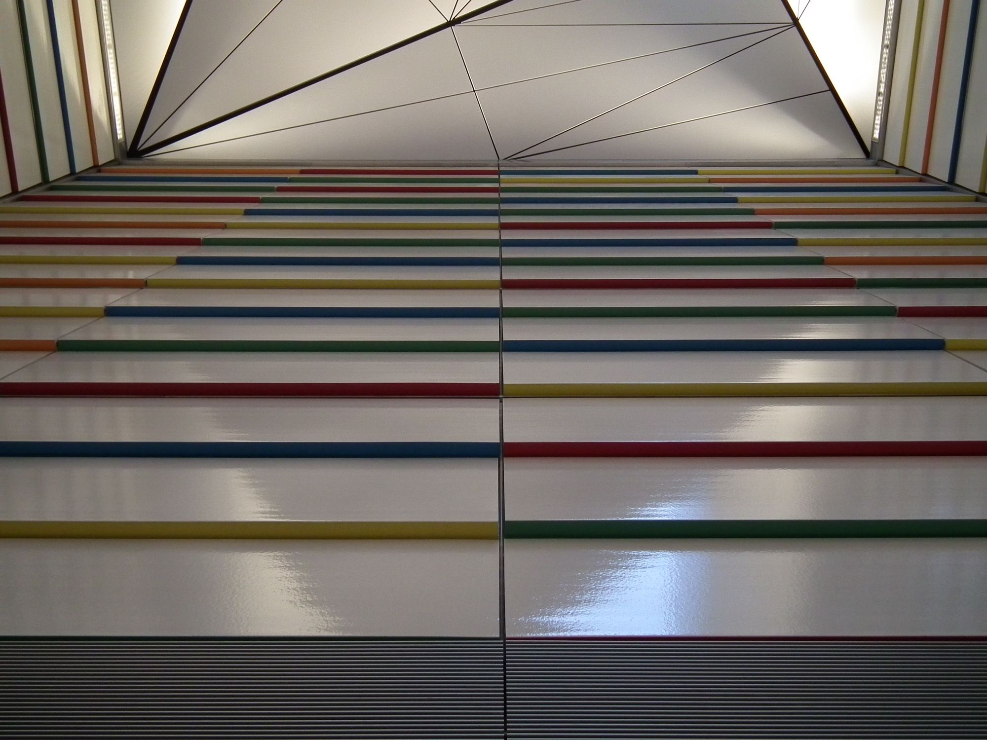 Looking up against a side wall. The familar rectangular wall panels here are plain white, but each one is angled such that the top is slightly further in than the bottom, and thus the lower face of each panel is visible — just 2-3 cm or so. These lower faces are various coloured red, green, yellow, and blue.