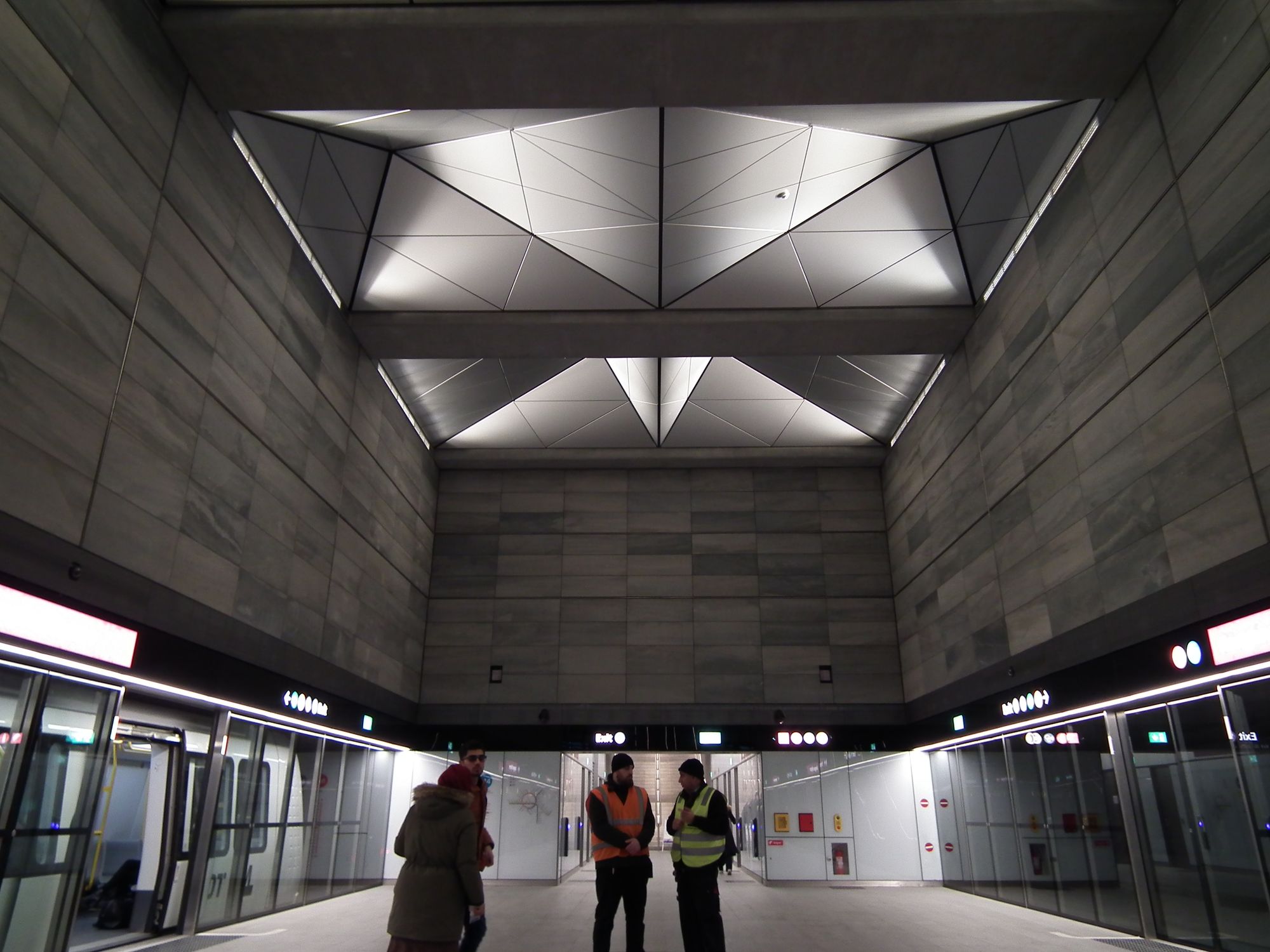 The platform area. There are a few people here. On the left and right, the glass safety walls, and on the left, a train is waiting. The ceiling is high, and includes two large fake skylights.