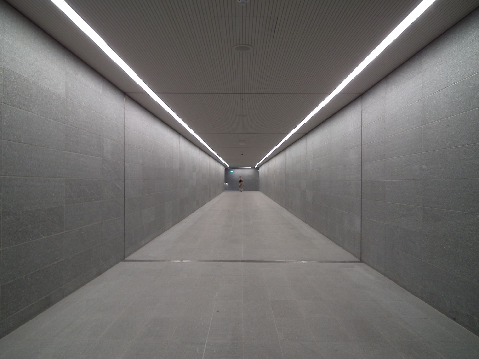 A long, plain, straight, doorless pedestrian tunnel. It is brightly lit. In the distance right at the end stands a person.
