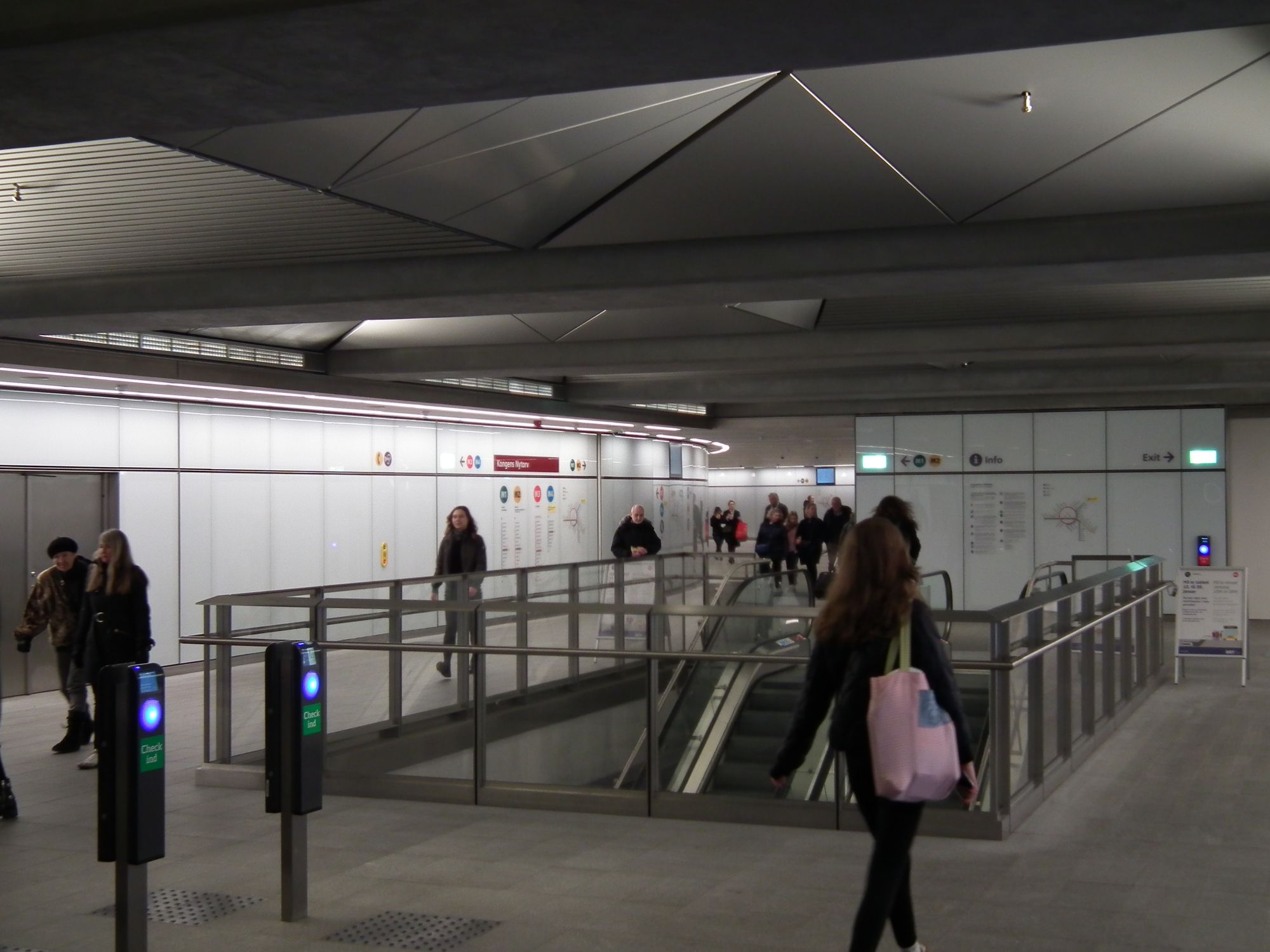 The underground ticket hall, with people coming and going. There are escalators down to the platform. The wall on the left curves away out of sight, towards the old M1/M2 lines.