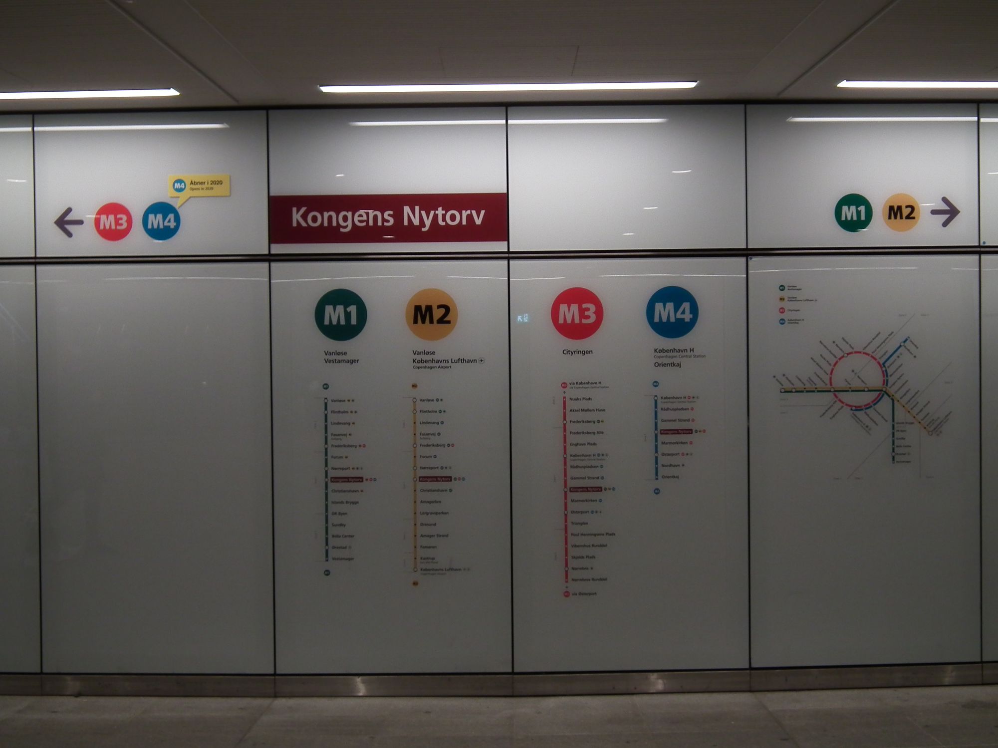 Part of the wall of the interchange area: the signage showing the station name, diagrams of the four lines that pass through here, schematic map, and arrows indicating left for M3/M4, right for M1/M2.