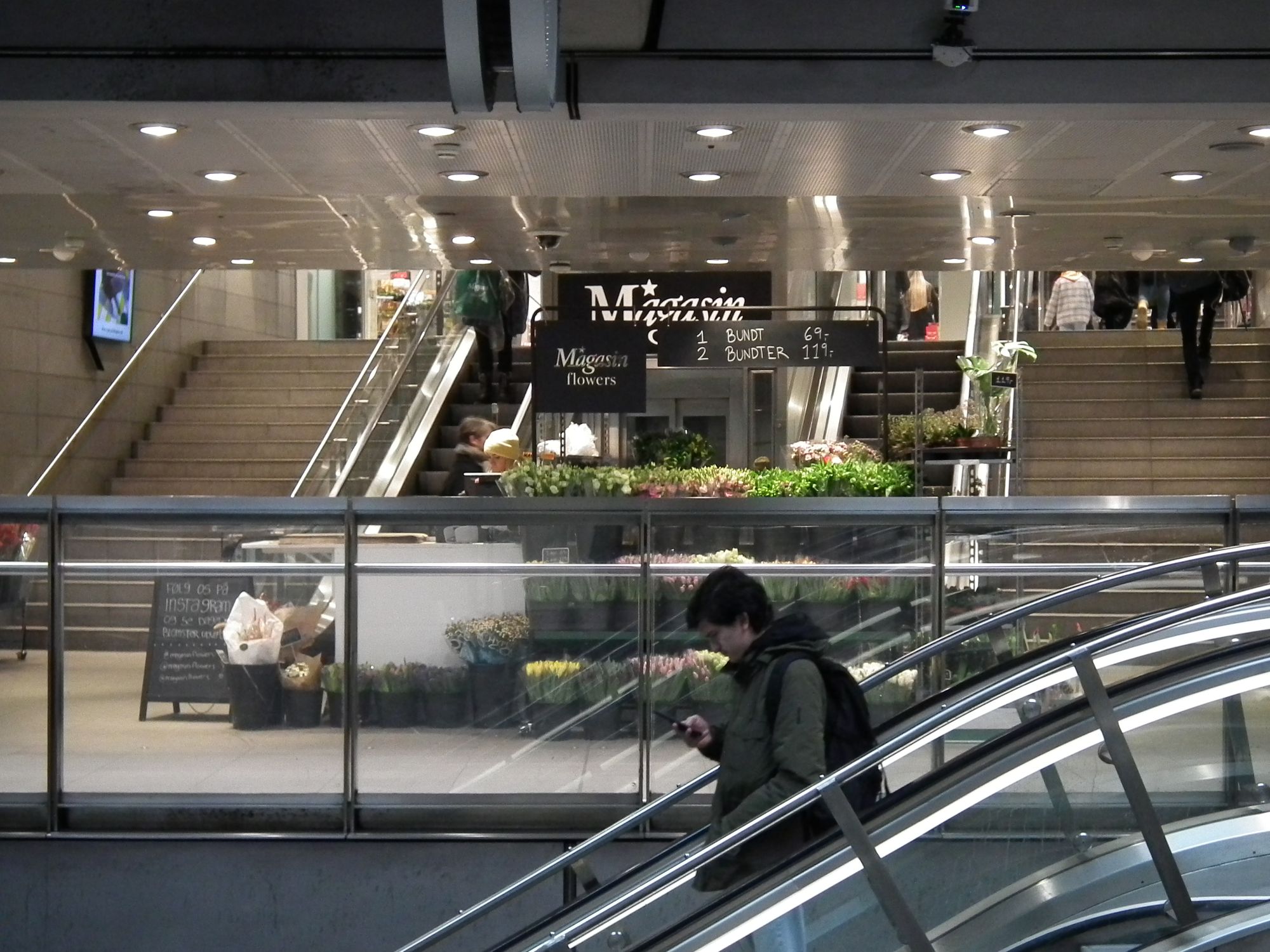 The sub-surface interchange level. In the foreground, an escalator descends (towards the M1/M2 lines); a person stands, travelling, looking at their phone. The focal point of the picture is the steps which lead directly up into 'Magasin', a department store. During opening hours, as now, the shop has a flower stall down here.