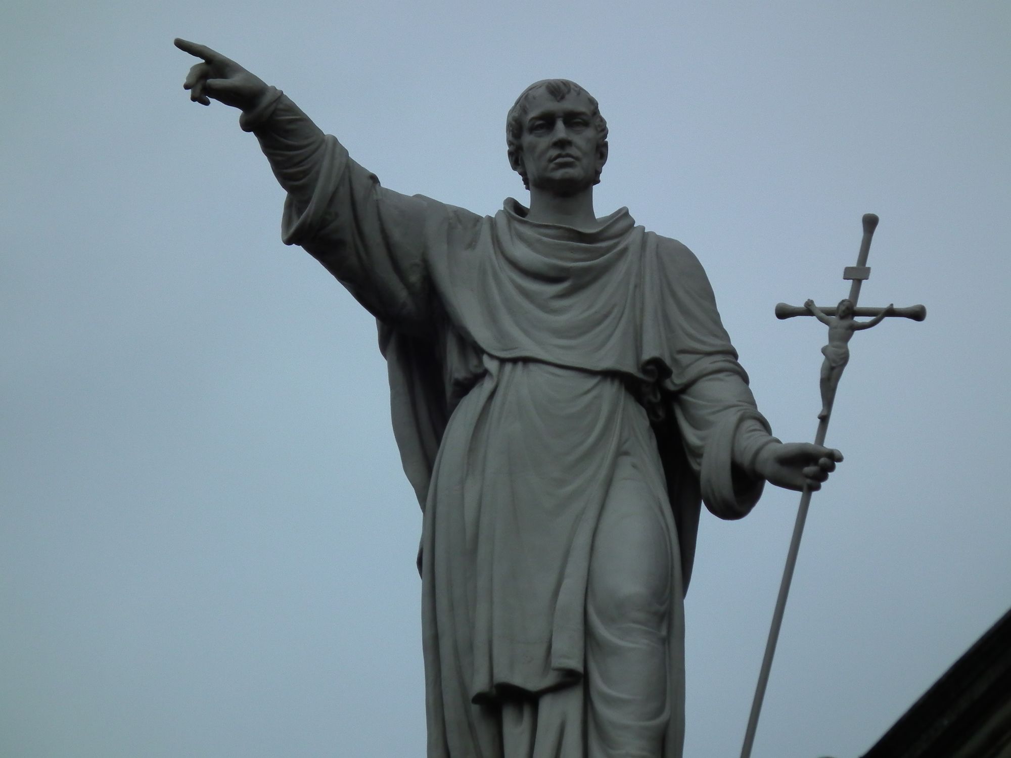 A closeup of one of the statues on top of the church. It's a man who holds some kind of cruciform object in his left hand. His right arm is raised, pointing off at ... something. Or nothing.