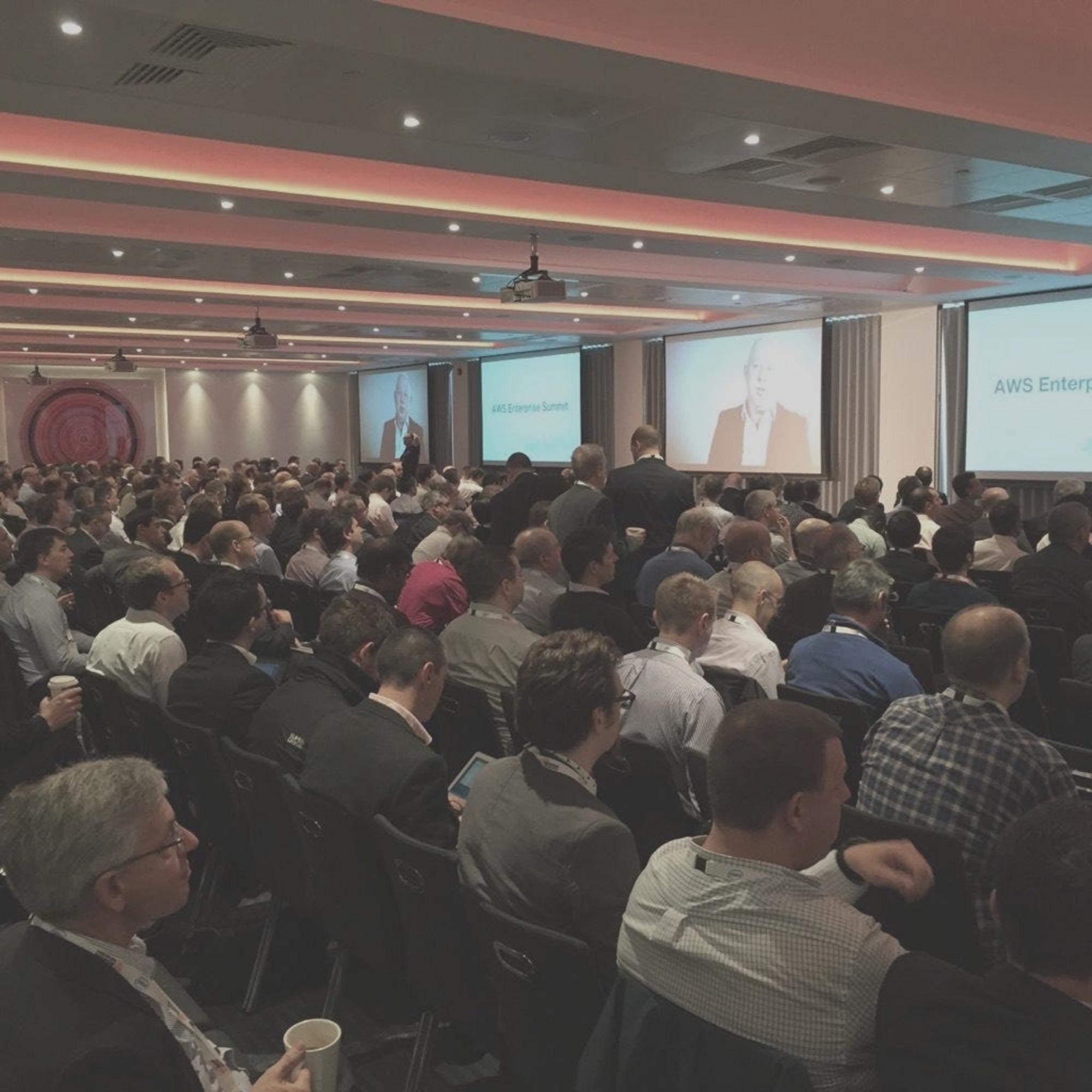 At the conference. The crowd sits, ready to hear the speakers. At the front of this low-ceilinged room is a series of large screens.