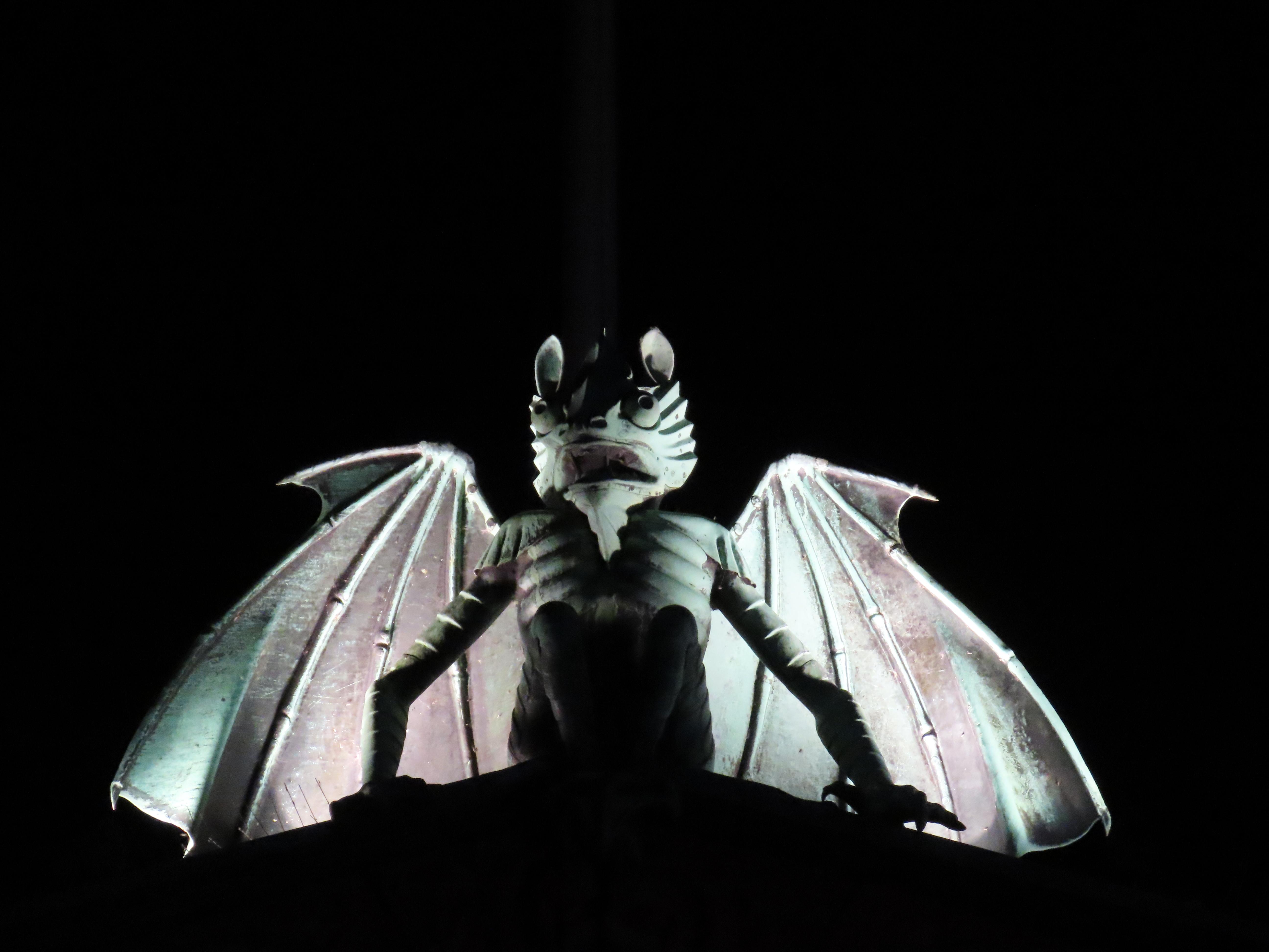 Night; atop the theatre building sits a large gargoyle, wings outspread, several meters wide. It is lit up from below, so all we see is the inky black night, and then this verdigris-tinted demon scowling over the city square.
