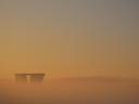 Two towers of a tall building peek out of a thick layer of mist. Apart from the towers, bottom left, the entire frame is a hazy orange, with a darker layer at the horizon, then becoming more green-blue higher in the sky.