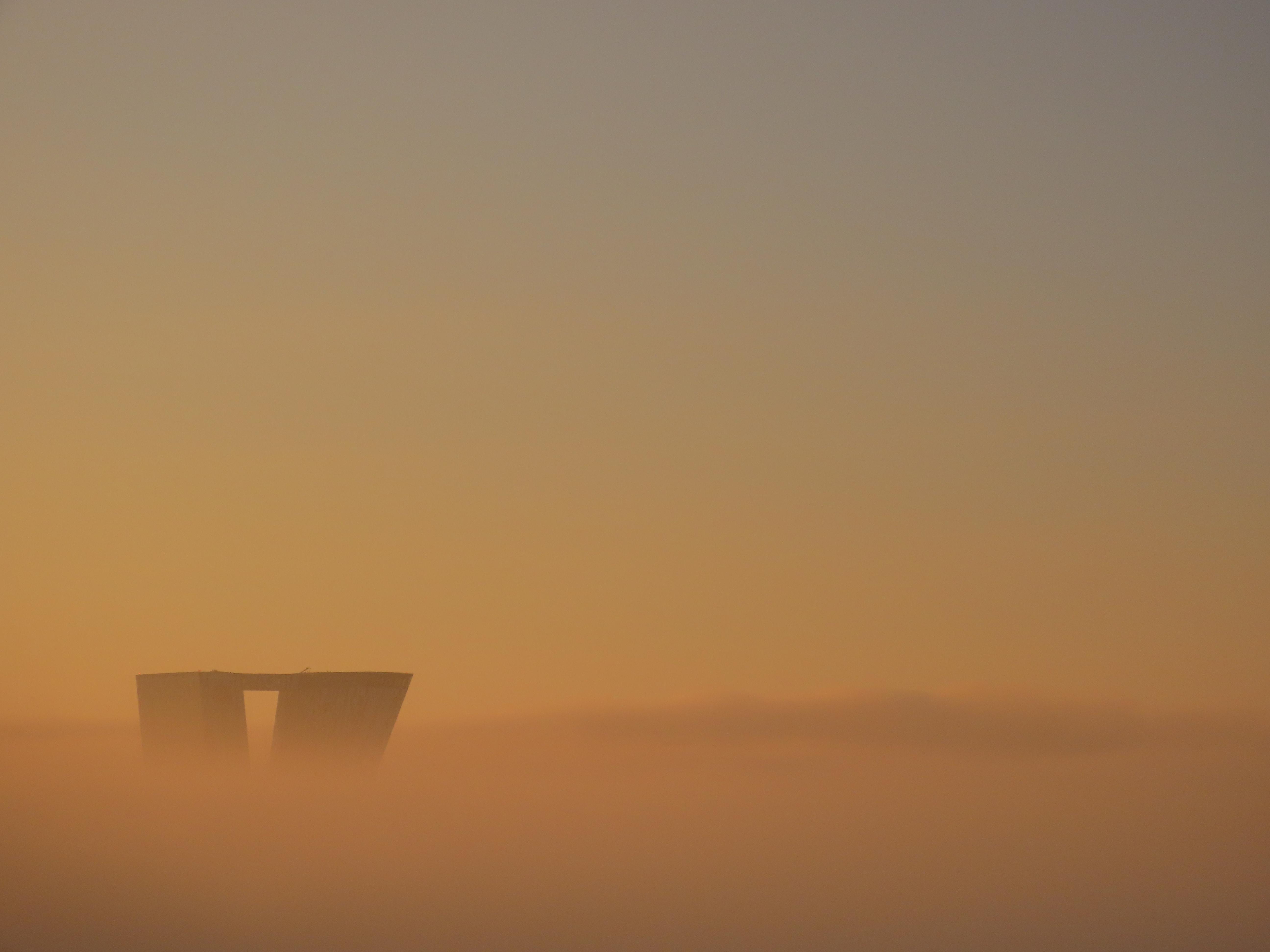 Two towers of a tall building peek out of a thick layer of mist. Apart from the towers, bottom left, the entire frame is a hazy orange, with a darker layer at the horizon, then becoming more green-blue higher in the sky.
