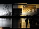 Aarhus harbour at night. Across the water, filling the upper half of the picture, is a warehouse; below that, the water, and the silhouettes of two people standing on the quayside, bottom right. On the side of the warehouse is (right) some banners advertising whatever, bathed in a golden light; and (left) a large mural, maybe 10 meters square, of a man with a weathered face, wearing an overcoat. He draws on a cigarette. He is looking towards the two silhouetted figures.