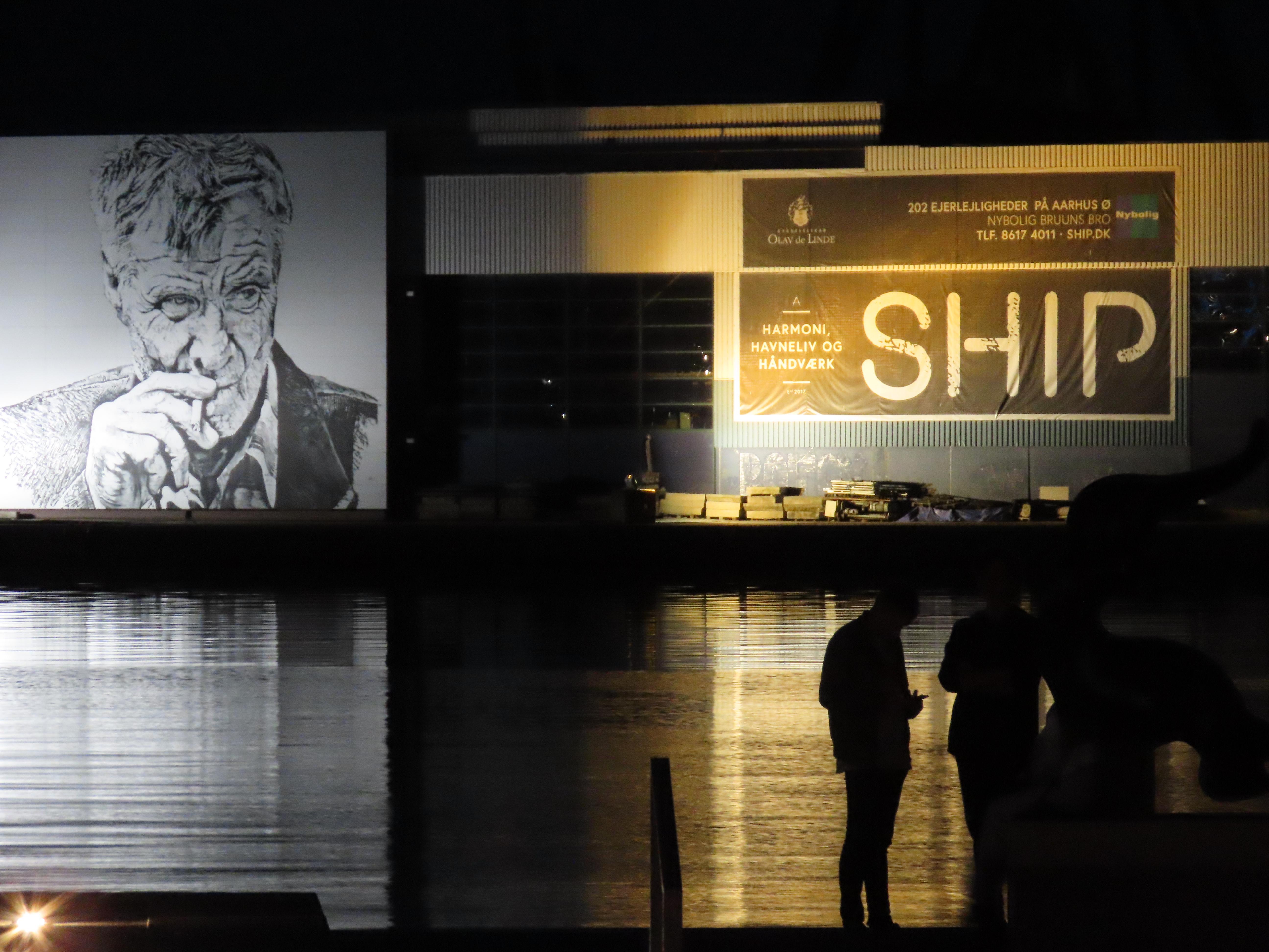 Aarhus harbour at night. Across the water, filling the upper half of the picture, is a warehouse; below that, the water, and the silhouettes of two people standing on the quayside, bottom right. On the side of the warehouse is (right) some banners advertising whatever, bathed in a golden light; and (left) a large mural, maybe 10 meters square, of a man with a weathered face, wearing an overcoat. He draws on a cigarette. He is looking towards the two silhouetted figures.