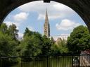 A church with a spire, on a sunny day with light cloud. In front of the church are deciduous trees, and then the river. The whole picture is framed above by a railway arch, making the church scene look a bit like it's in a snow globe.