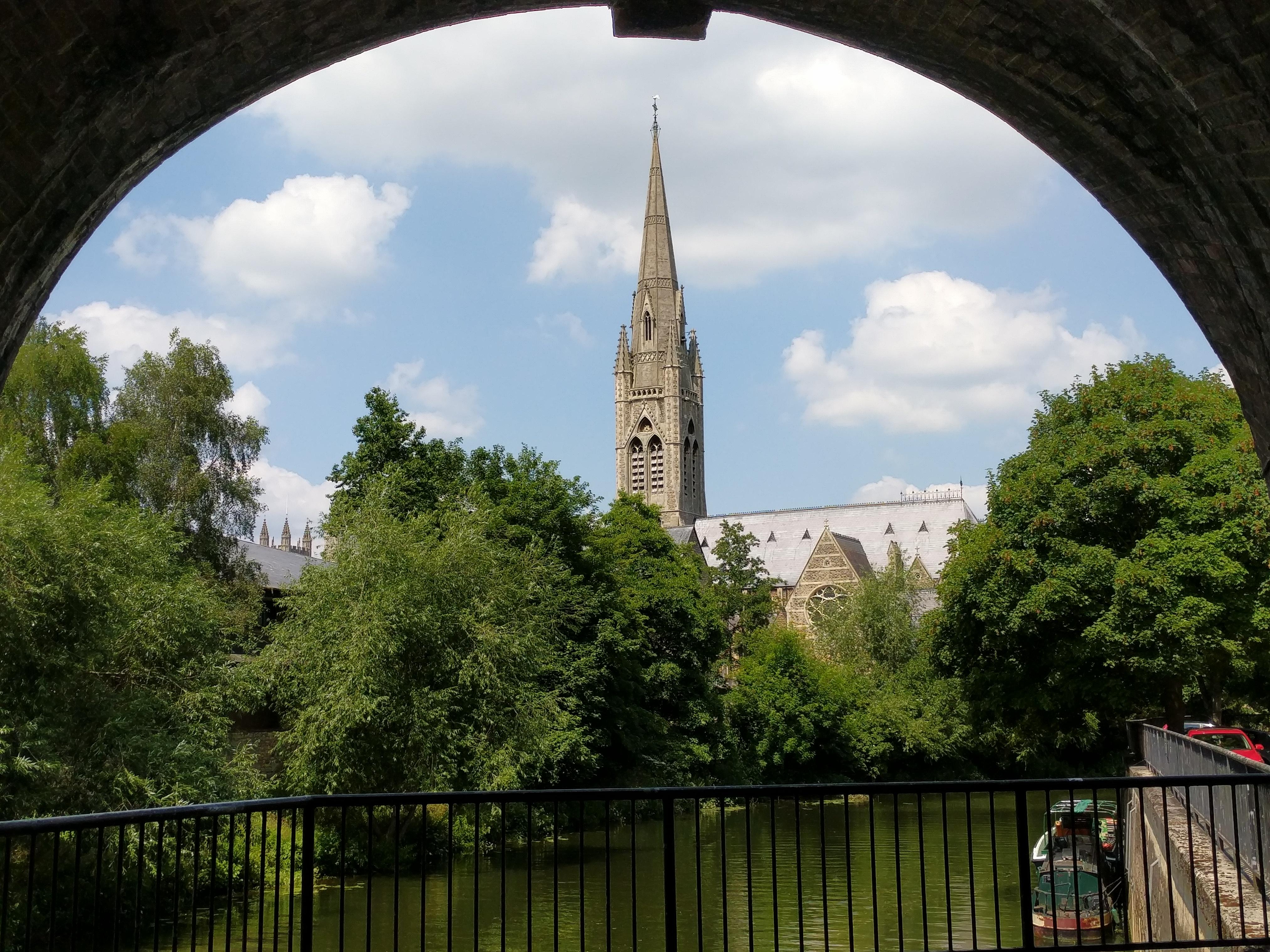 A church with a spire, on a sunny day with light cloud. In front of the church are deciduous trees, and then the river. The whole picture is framed above by a railway arch, making the church scene look a bit like it's in a snow globe.