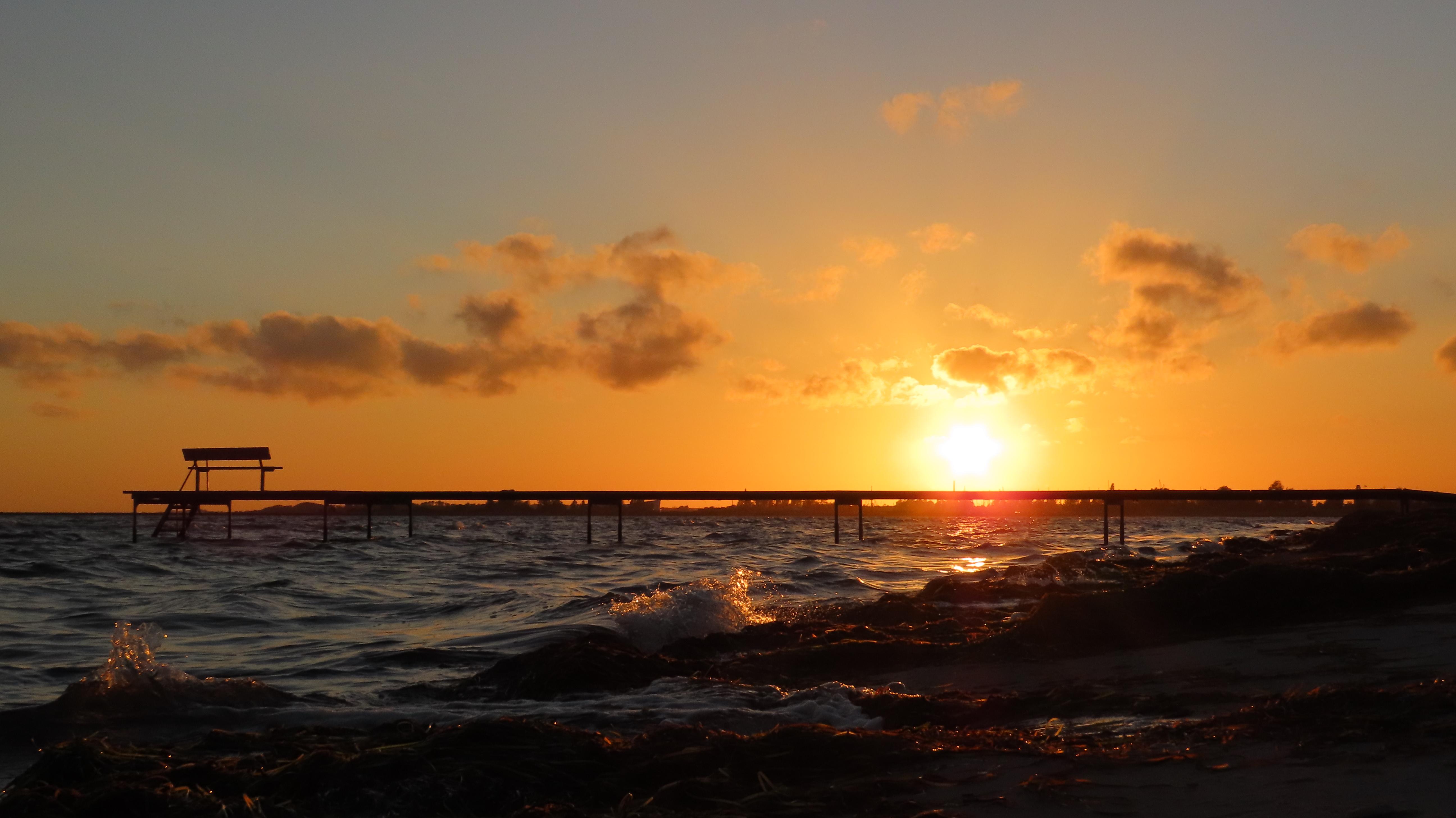 A colourful sunset at the beach. There is a jetty going some 25 meters out to sea, with a bench at the end.