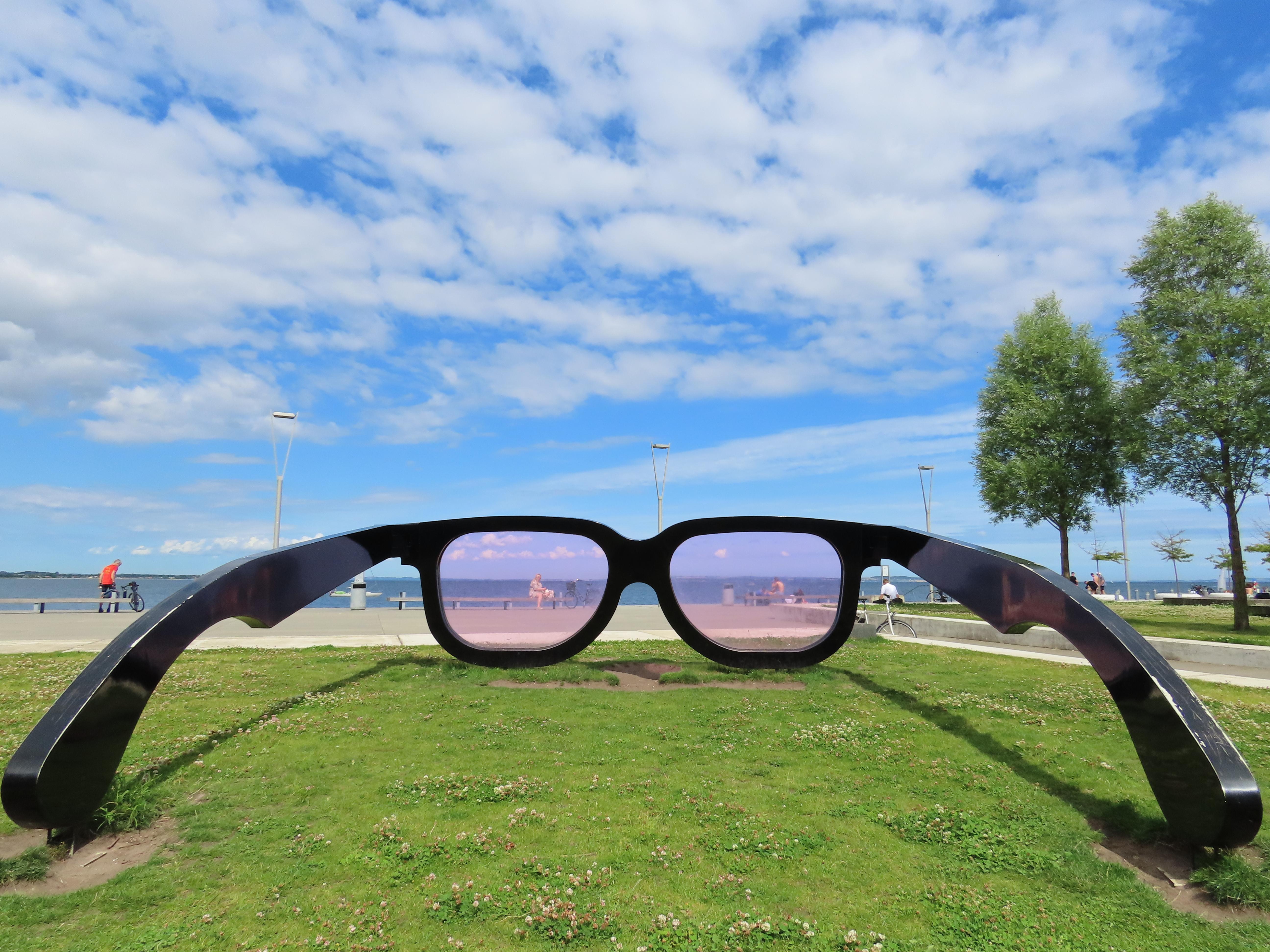 A park, next to the sea, on a sunny day with light cloud. On the grassy area is a large sculpture: a huge pair of glasses, arms opened, forming three sides of a square, maybe 3.5 meters per side. The lenses have a subtle pink tinge. Through the left lens, we see a woman sat on a bench by the water.